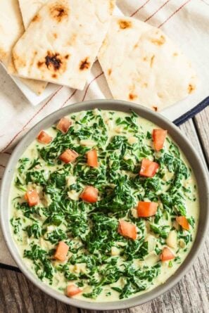 A bowl of Indian Creamed Spinach garnished with diced tomatoes, served with pieces of naan on a cloth.