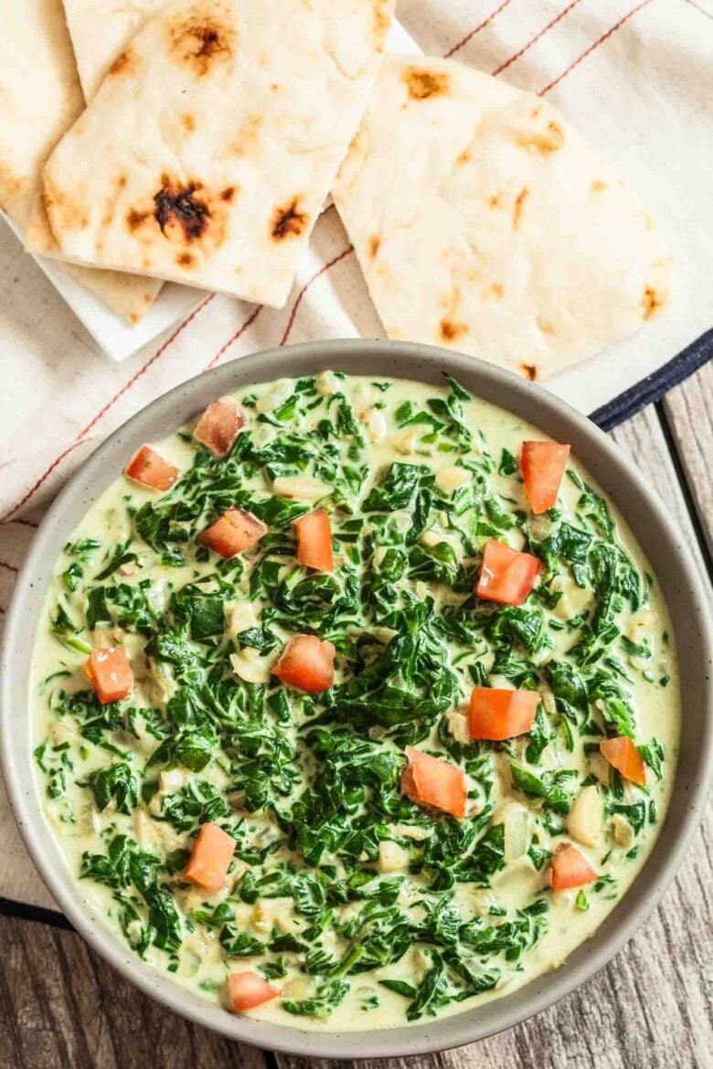 A bowl of Indian Creamed Spinach garnished with diced tomatoes, served with pieces of naan on a cloth.