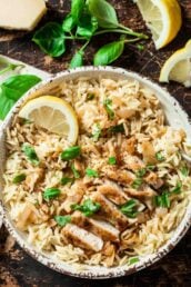 A bowl of seasoned orzo topped with sliced grilled chicken, fresh basil leaves, and a lemon wedge to serve lemon chicken orzo at home.