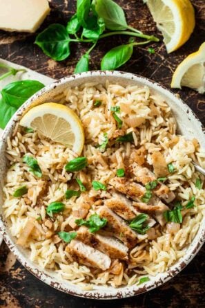 A bowl of seasoned orzo topped with sliced grilled chicken, fresh basil leaves, and a lemon wedge to serve lemon chicken orzo at home.