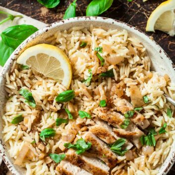 A bowl of seasoned orzo topped with sliced grilled chicken, fresh basil leaves, and a lemon wedge to serve lemon chicken orzo at home.