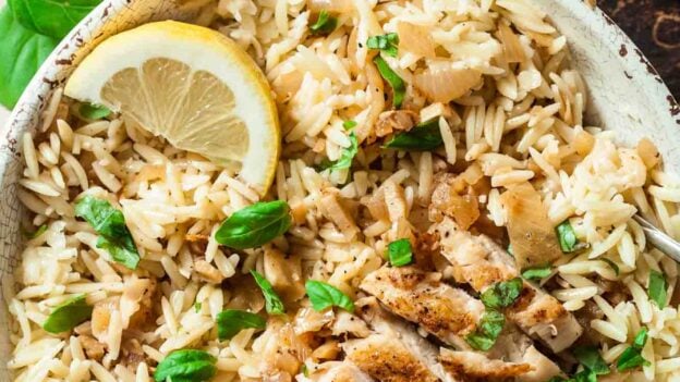 A bowl of seasoned orzo topped with sliced grilled chicken, fresh basil leaves, and a lemon wedge to serve lemon chicken orzo at home.
