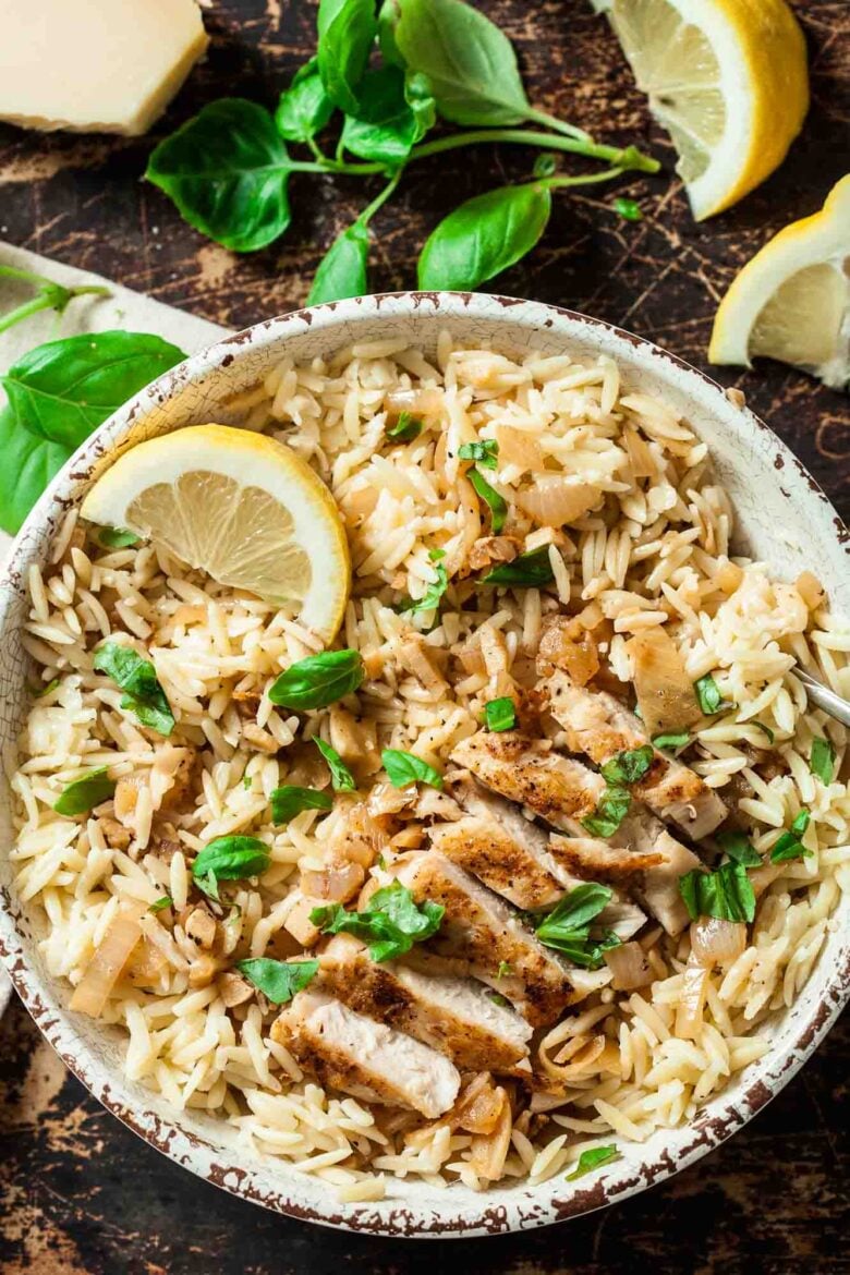 A bowl of seasoned orzo topped with sliced grilled chicken, fresh basil leaves, and a lemon wedge to serve lemon chicken orzo at home.