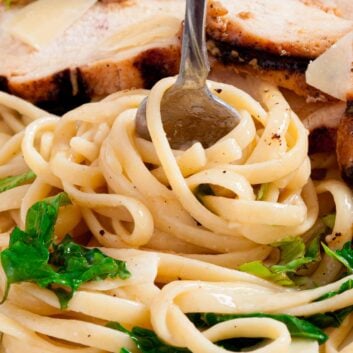Close-up of lemon chicken pasta with leafy greens and sliced grilled chicken, topped with parmesan cheese, with a fork twirling the pasta.