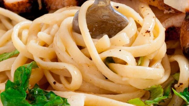 Close-up of lemon chicken pasta with leafy greens and sliced grilled chicken, topped with parmesan cheese, with a fork twirling the pasta.