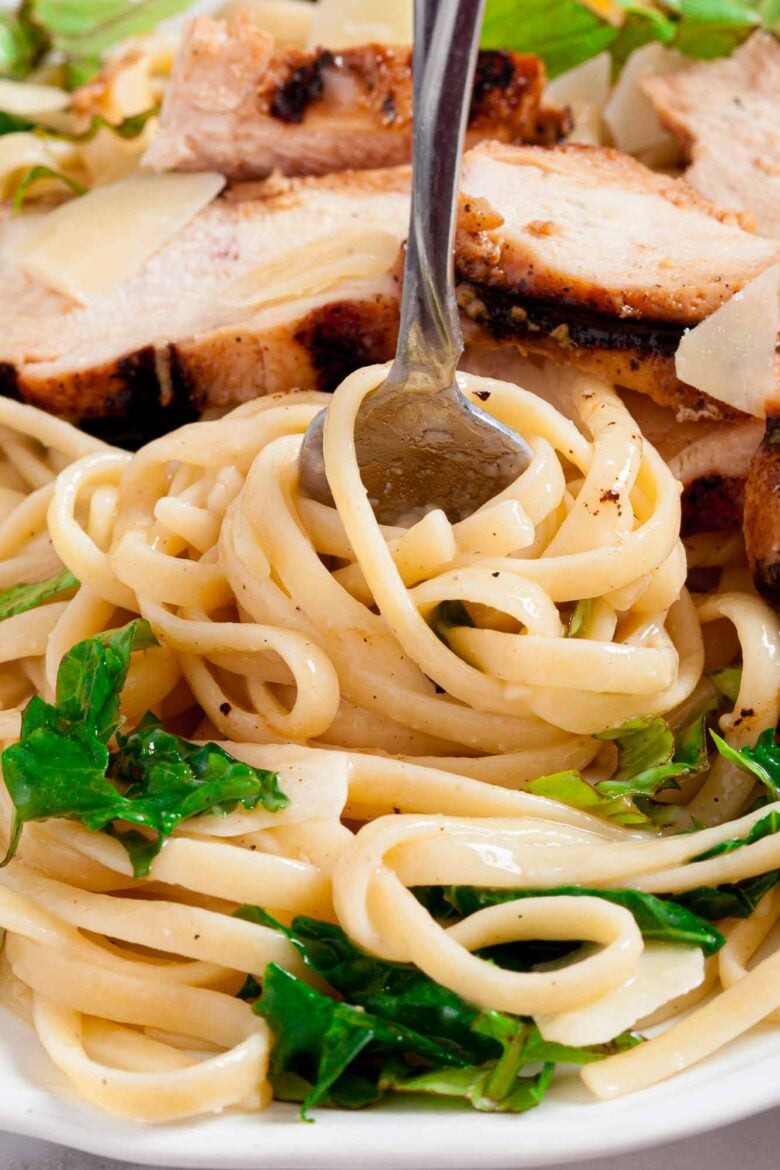 Close-up of lemon chicken pasta with leafy greens and sliced grilled chicken, topped with parmesan cheese, with a fork twirling the pasta.
