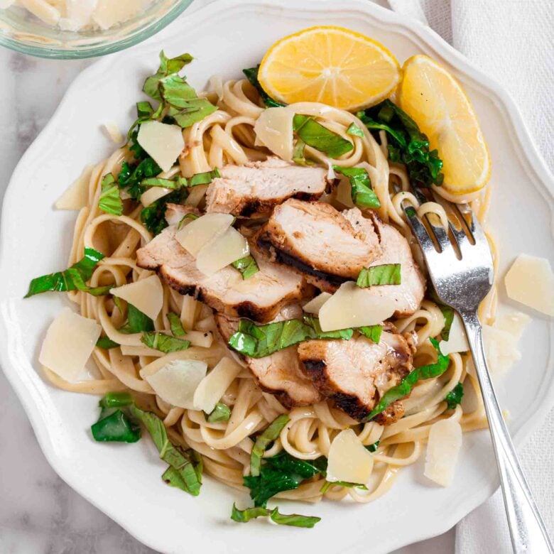White plate with linguine pasta topped with grilled chicken slices, fresh spinach, shaved parmesan, and lemon wedges; a fork rests on the side.