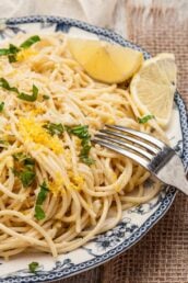 A plate of spaghetti al limone garnished with grated cheese, chopped herbs, lemon zest, and lemon wedges, with a fork resting on the plate.