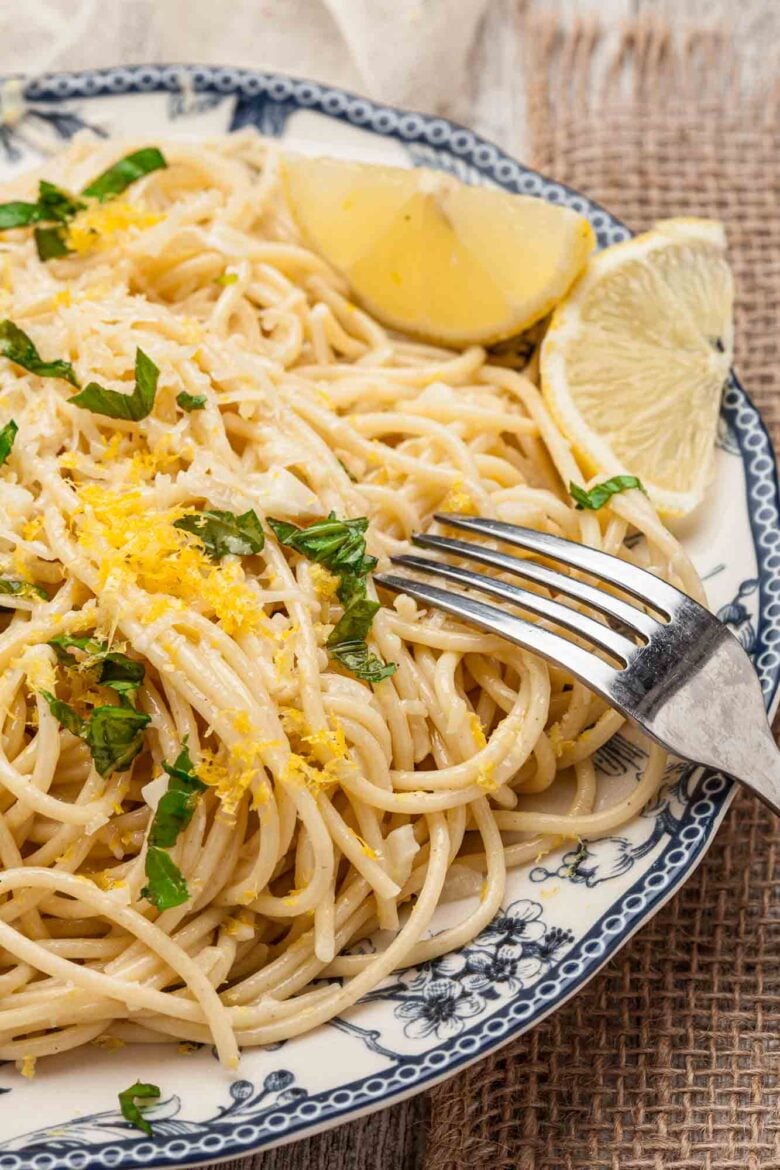 A plate of spaghetti al limone garnished with grated cheese, chopped herbs, lemon zest, and lemon wedges, with a fork resting on the plate.
