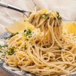 A plate of spaghetti al limone topped with grated lemon zest and herbs, with a fork lifting a portion. Two lemon wedges are visible on the side.