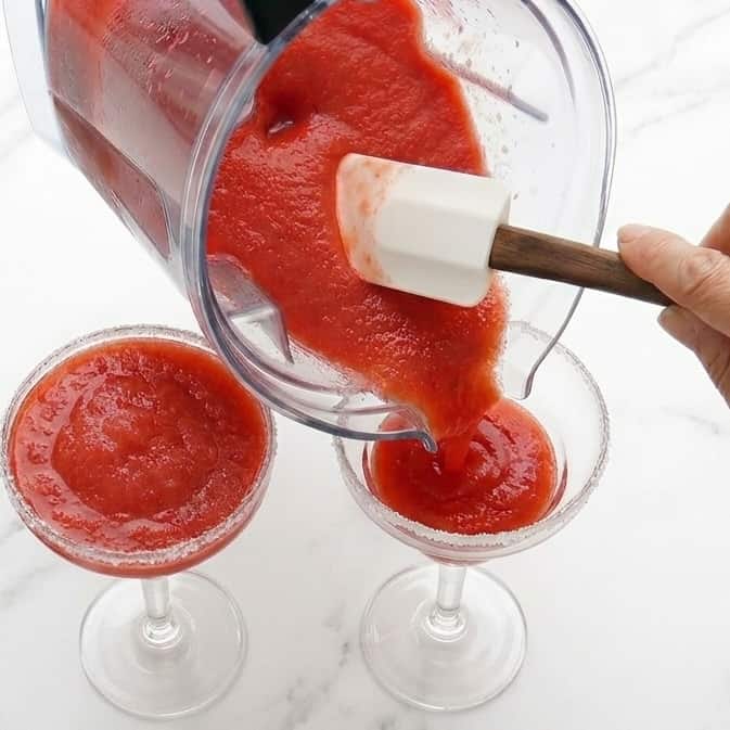 A person pours a 4-ingredient strawberry daiquiri, made with frozen strawberries, from a blender into two sugar-rimmed cocktail glasses using a spatula.
