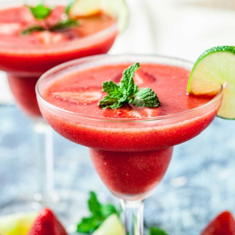 A 4-ingredient Strawberry Daiquiri, made with frozen strawberries, garnished with fresh mint leaves and a lime wedge in a margarita glass, with strawberries and lime pieces in the background.
