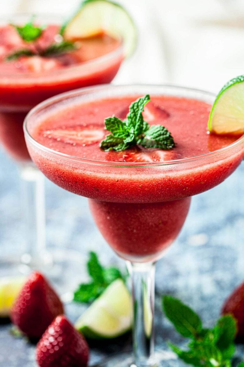 A 4-ingredient Strawberry Daiquiri, made with frozen strawberries, garnished with fresh mint leaves and a lime wedge in a margarita glass, with strawberries and lime pieces in the background.