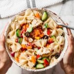 A bowl of easy pasta salad with bowtie pasta, sliced cucumbers, black olives, diced bell peppers, and bits of bacon on a marble surface with a striped towel nearby.