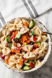 A bowl of easy pasta salad with bowtie pasta, sliced cucumbers, black olives, diced bell peppers, and bits of bacon on a marble surface with a striped towel nearby.