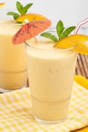 Two clear glasses full of mango smoothie with straws and umbrellas coming out of them.