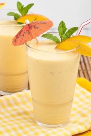 Two clear glasses full of mango smoothie with straws and umbrellas coming out of them.