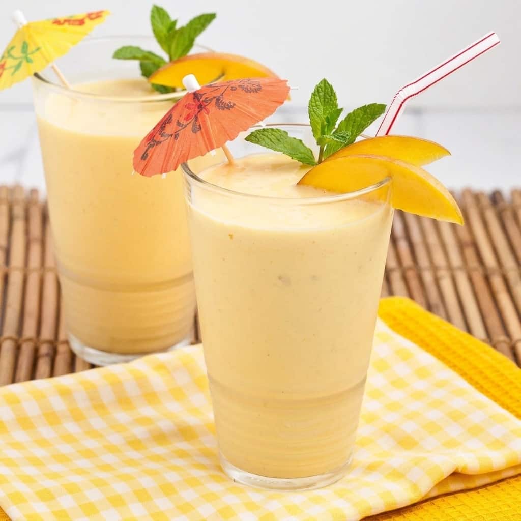 Two clear glasses full of mango smoothie with straws and umbrellas coming out of them.