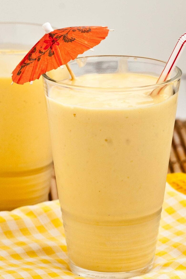 Tall clear glass filled with Mango smoothie with a fun drink umbrella and straw.