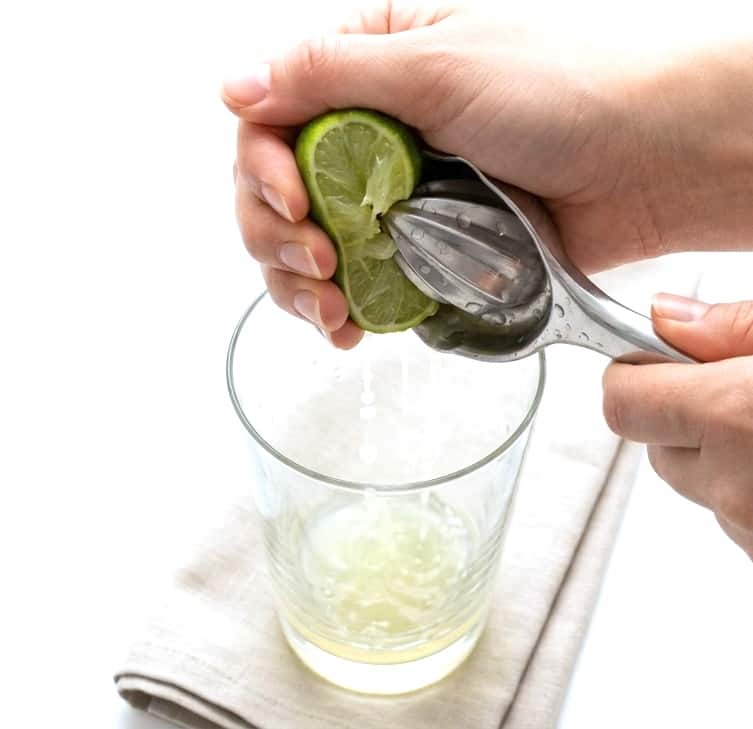 Hand holding half a lime and juicing the fresh lime juice into a tall highball glass. 