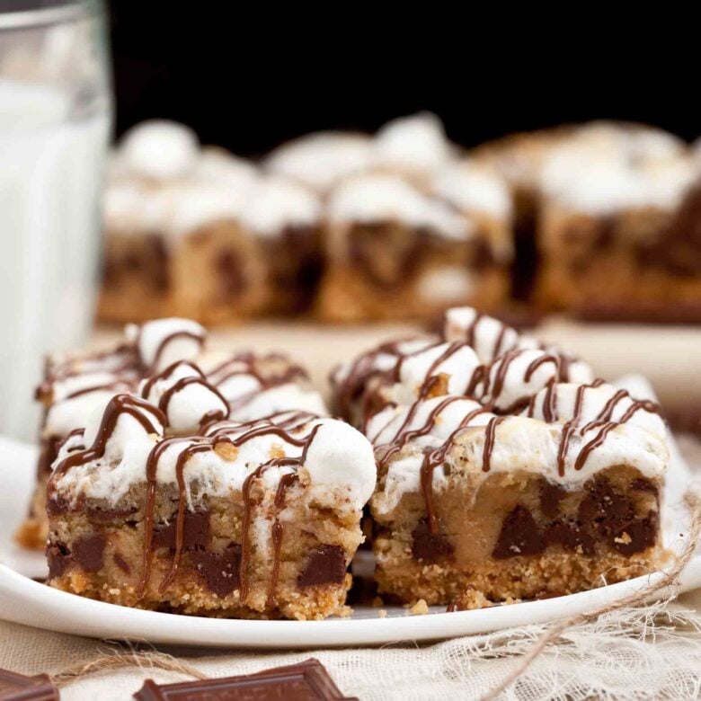 A plate of s'mores dessert bars with chocolate chips, marshmallows, and chocolate drizzle, perfect for summer gatherings, with a glass of milk in the background.
