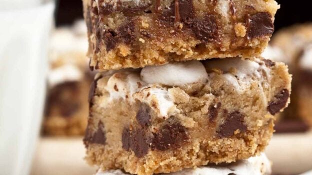 Three stacked s'mores dessert bars topped with marshmallows and chocolate drizzle, on a white plate with chocolate pieces and a glass of milk in the background.