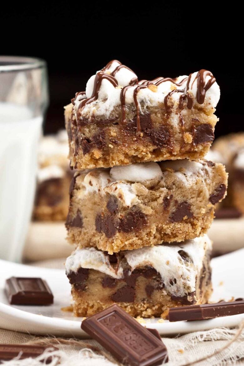 Three stacked s'mores dessert bars topped with marshmallows and chocolate drizzle, on a white plate with chocolate pieces and a glass of milk in the background.