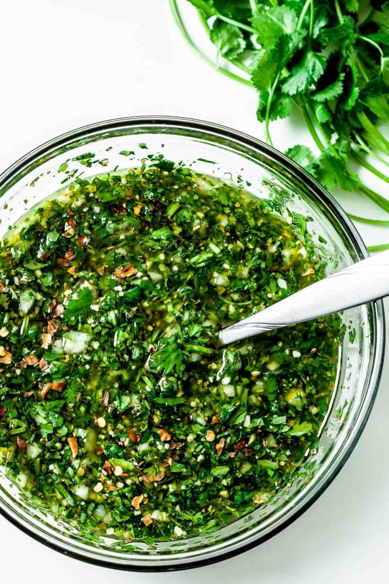 Clear bowl of chimichurri sauce stirred and mixed with a spoon in it for serving.