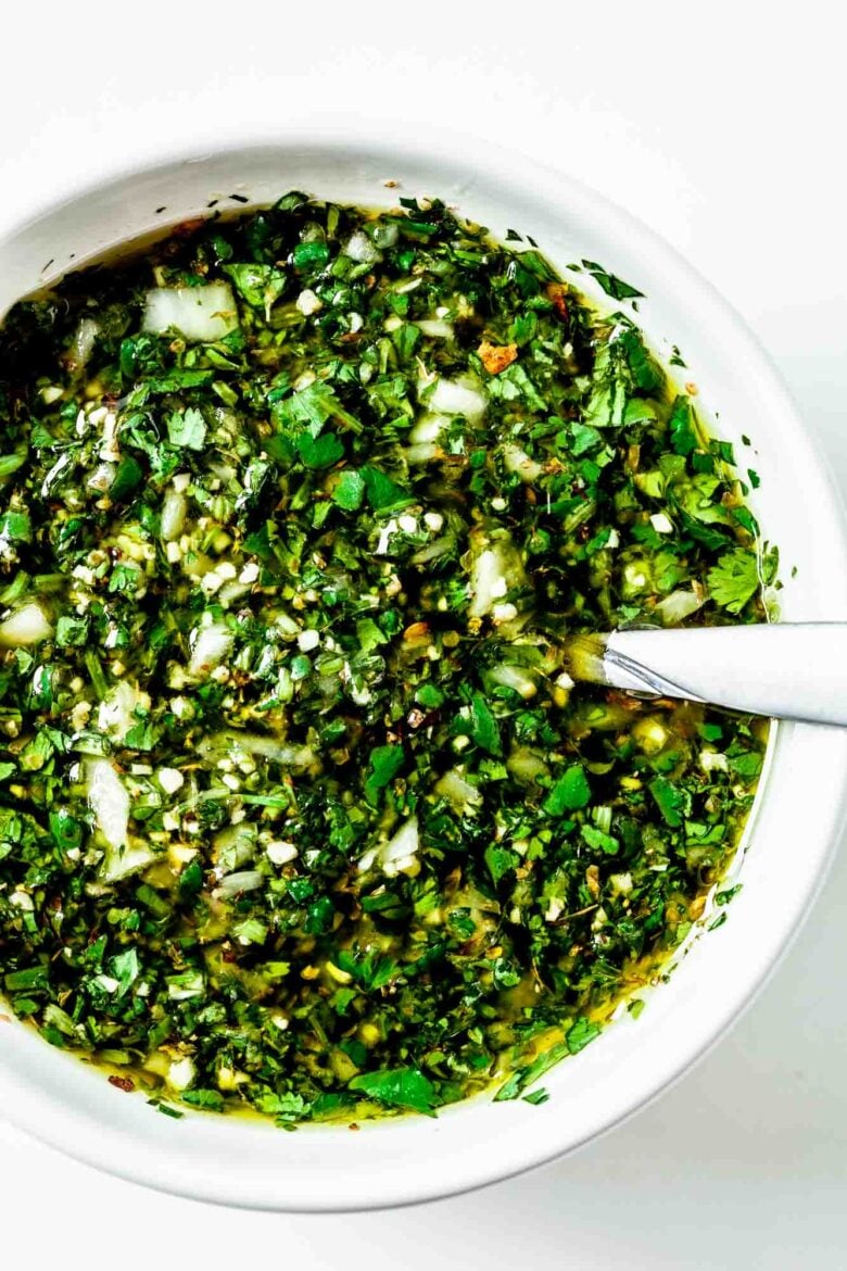 White bowl full of Chimichurri sauce all mixed together with a spoon in it for serving.