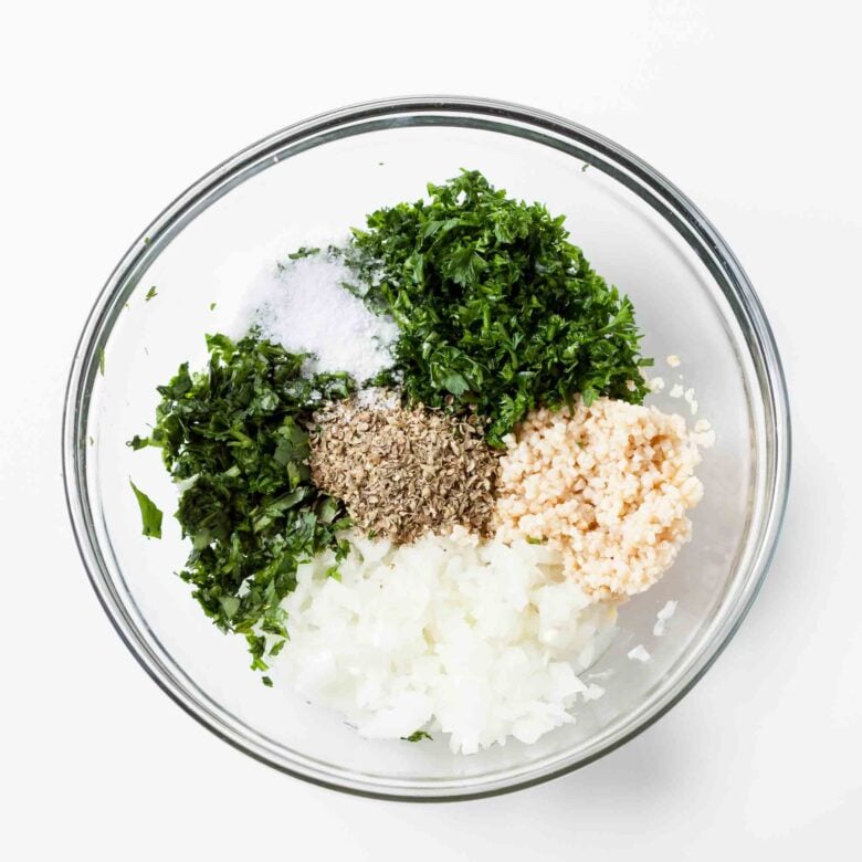 Adding all the ingredients needed to make chimichurri sauce in a large clear glass mixing bowl.