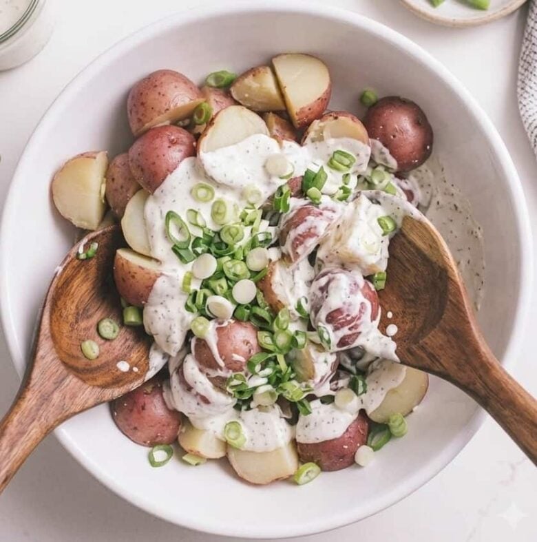 Boiled potatoes in a white bowl with the Ranch salad dressing on top and two wooden spoons ready to stir.