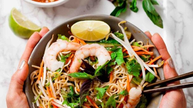 Hands holding a serving bowl full of thin rice noodles and jump shrimp to create a spring roll bowl.