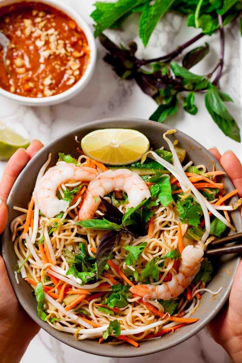 Hands holding a spring roll filled with thin rice noodles, fresh veggies, and jumbo shrimp in a serving bowl.
