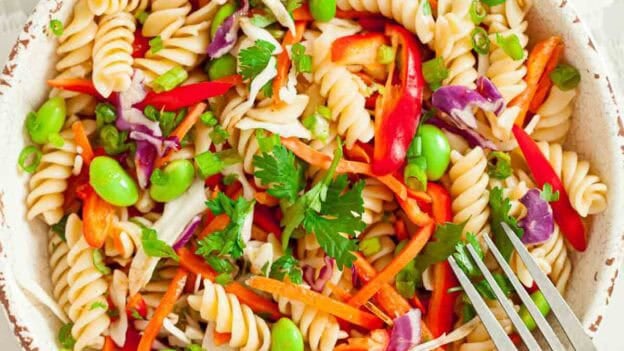 White bowl full of cooked asian pasta salad with a silver fork getting ready to take a scoop.