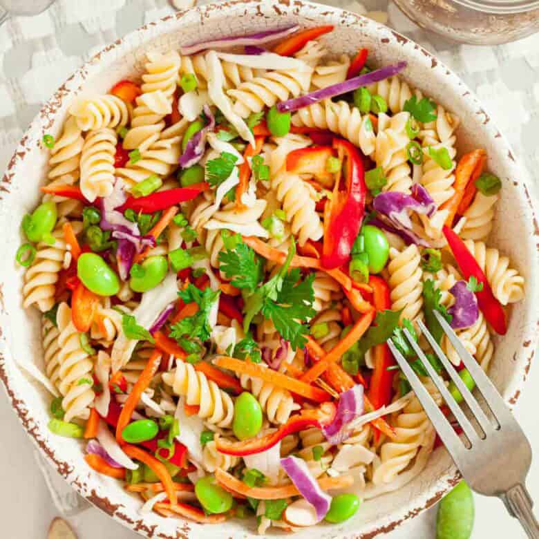 White bowl full of cooked asian pasta salad with a silver fork getting ready to take a scoop.