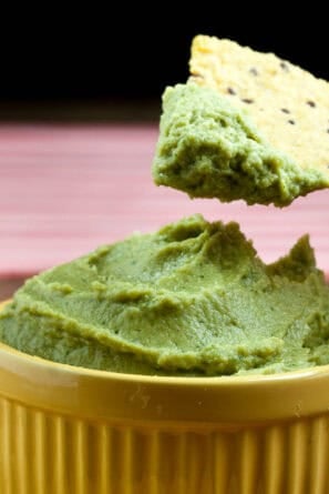 Hand holding tortilla chip and dipping it into a yellow bowl full of avocado hummus.