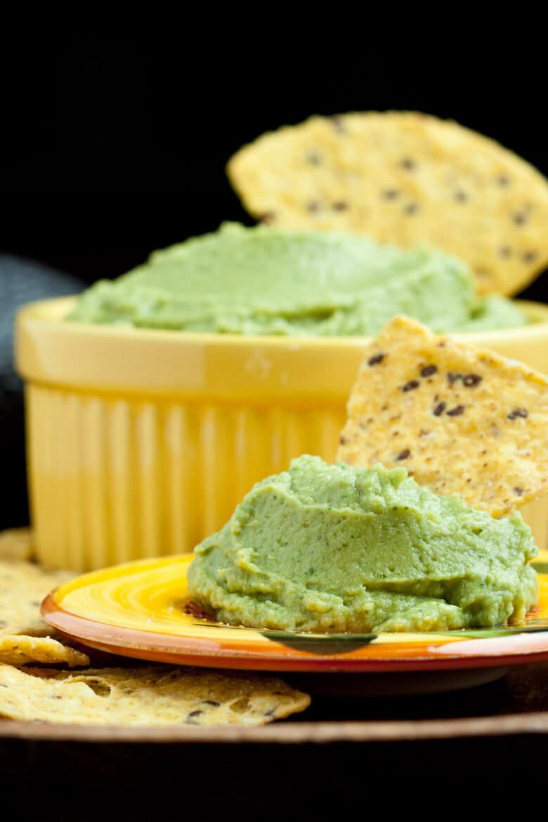 Chip dipping into avocado hummus and a small yellow bowl of hummus sitting in the background.