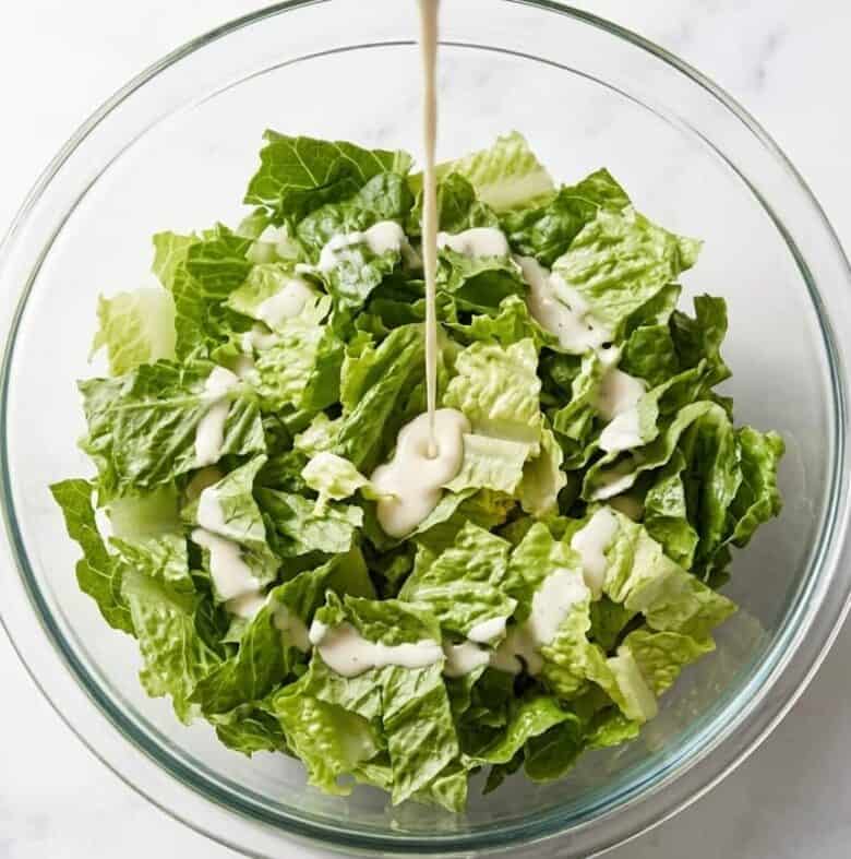 Pouring the Caesar dressing over the shredded lettuce in a glass bowl.