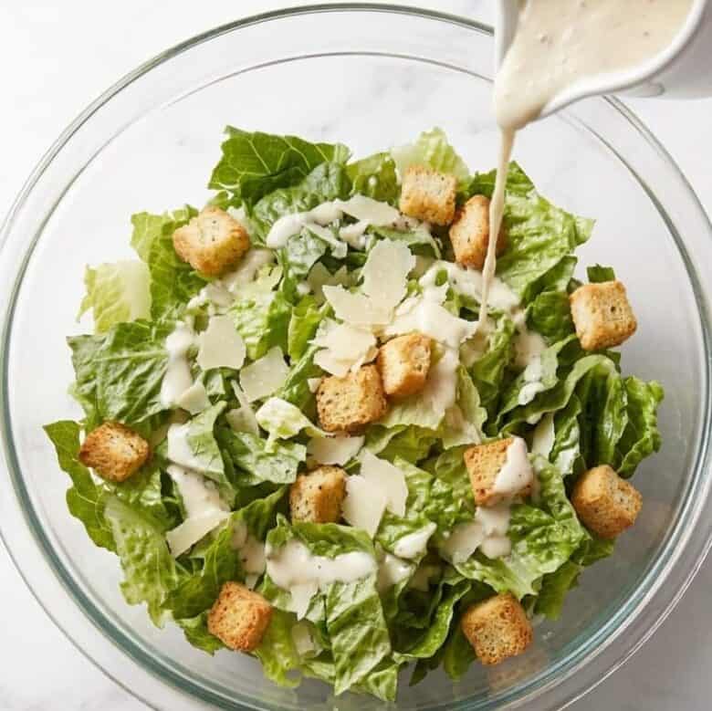 Clear glass bowl full of Caesar Salad with dressing being added.