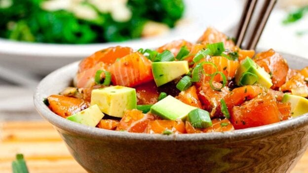 Brown bowl full of salmon poke bowl with brown chopsticks sticking out.