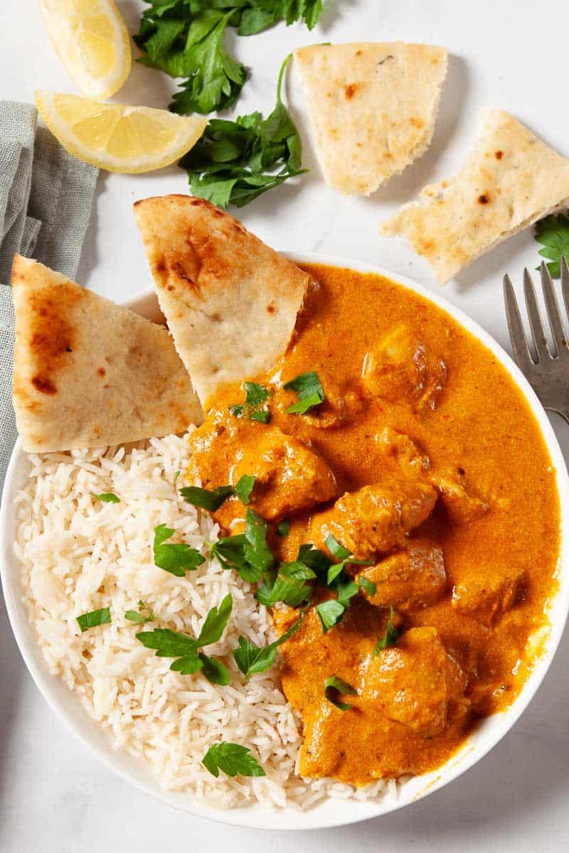 Overhead view looking down on Butter Chicken in a white serving bowl with rice and naan bread. 