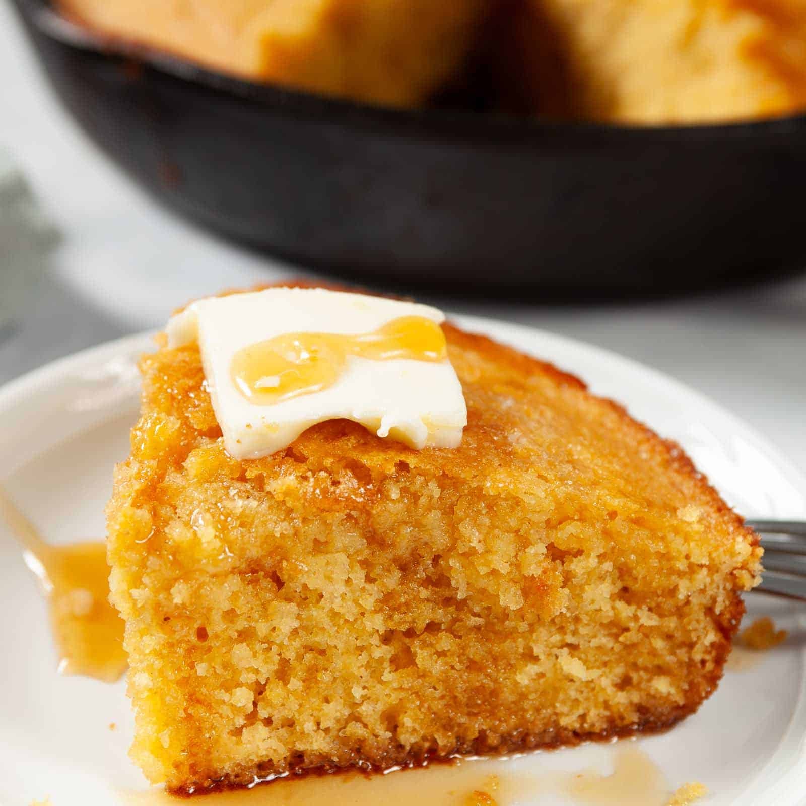 Close up of Honey Cornbread on a white plate topped with a cube of butter and a drizzle of honey.