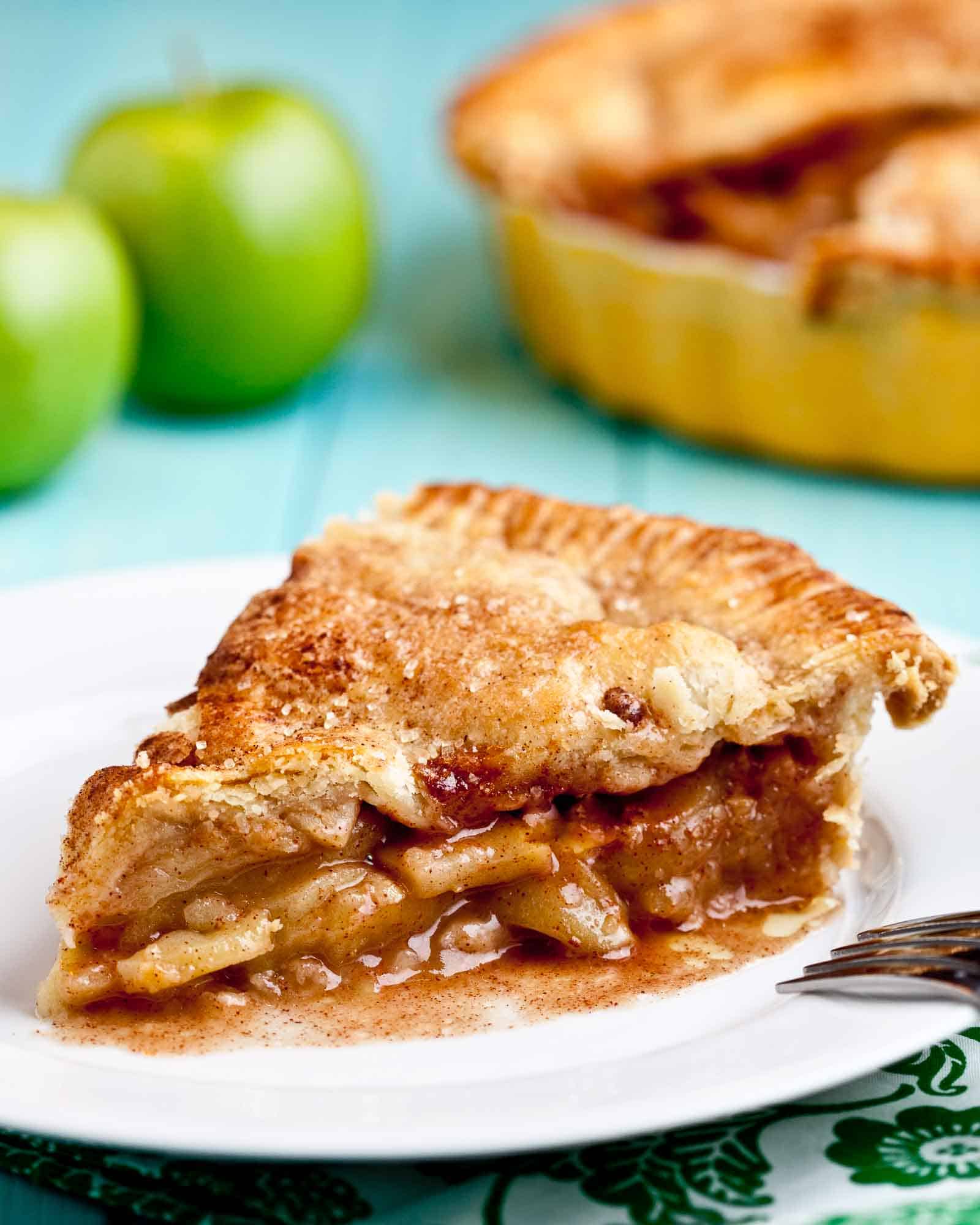 Apple pie with flaky butter crust sliced on a white plate with green apples in background. 